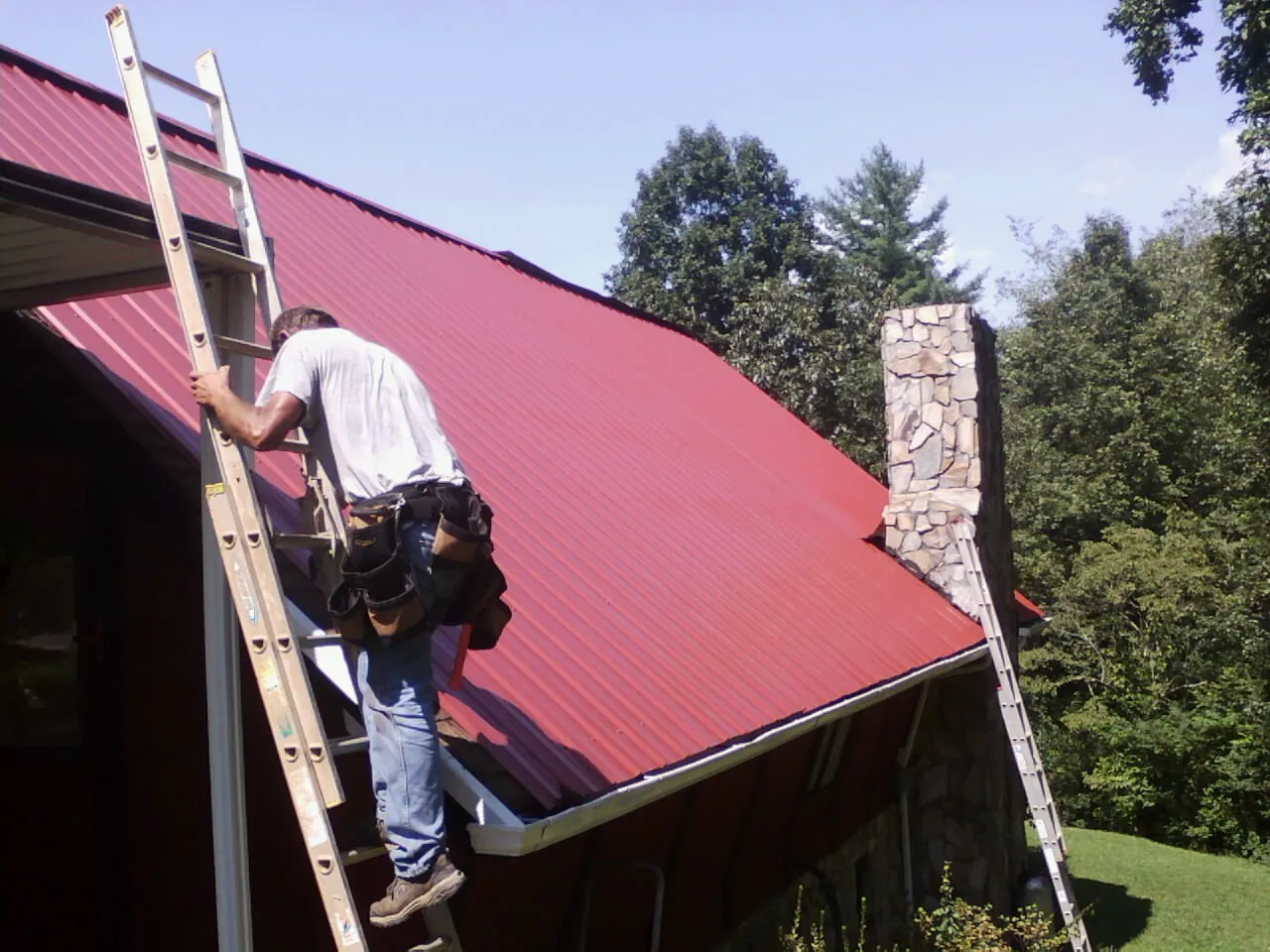 Roofer on a ladder beside a red metal roof and stone chimney — Soffit & Fascia Repair by Roofing Brothers in Westborough